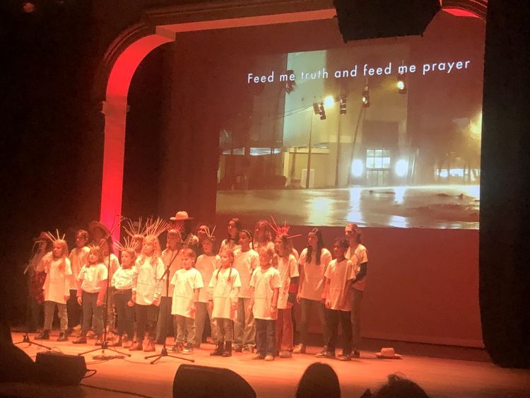 A group of children and young people wearing headdresses and singing on a stage with a large video projected behind them
